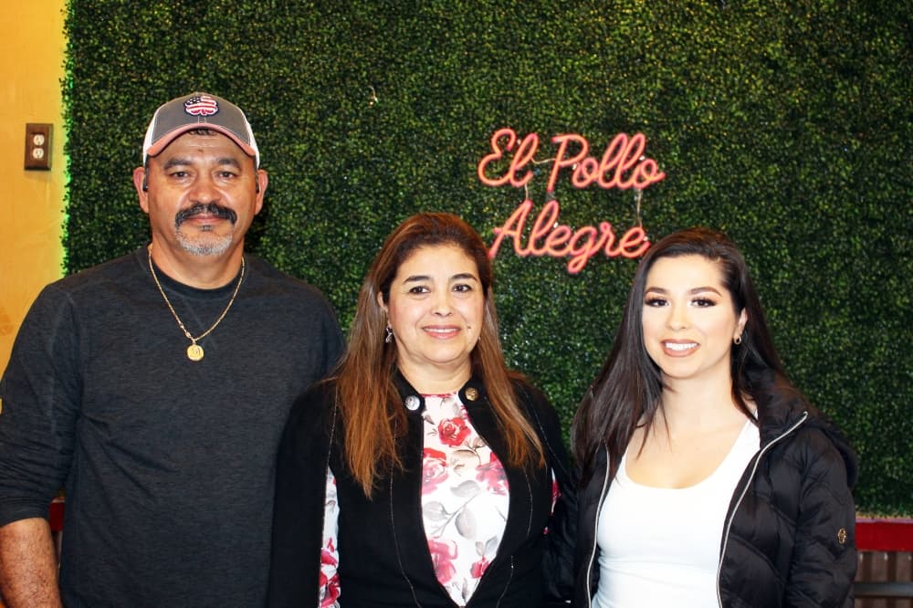 One male and two females standing in front of a wall with El Pollo Alegre signage. 