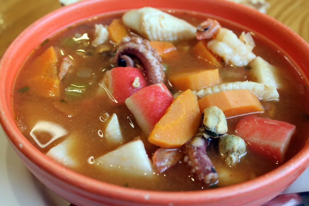 Soup in a bowl with fish, shrimp and octopus in a red/brown broth. 