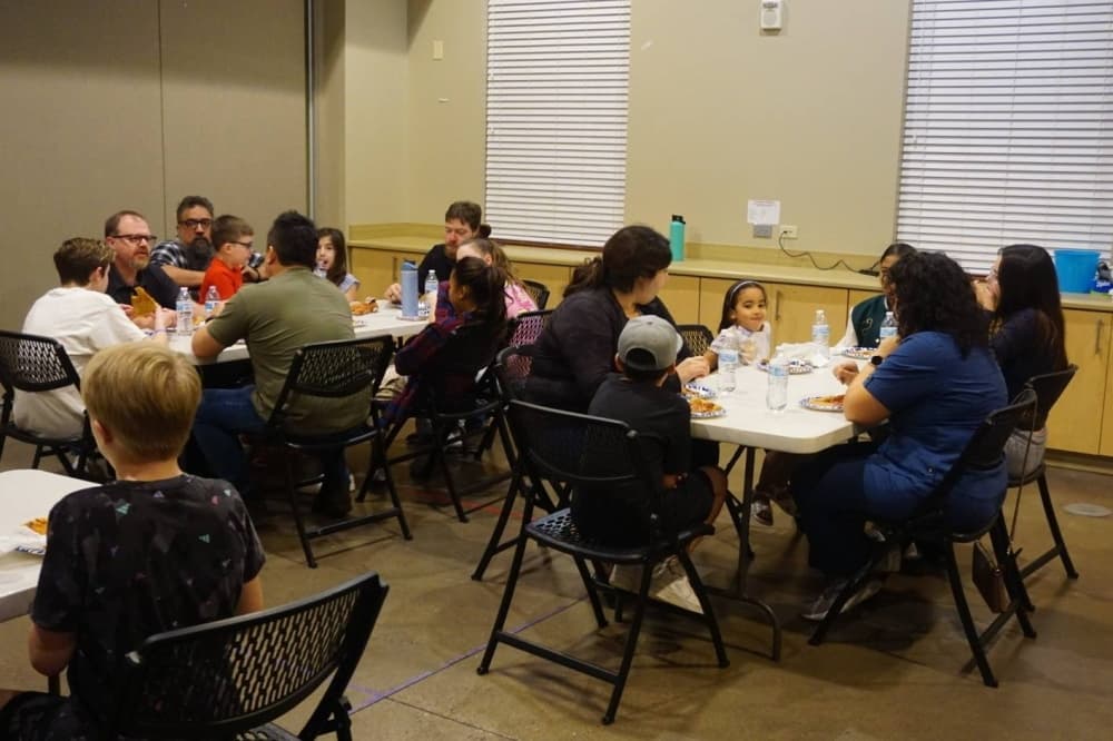 People eating pizza around three tables in a room. 