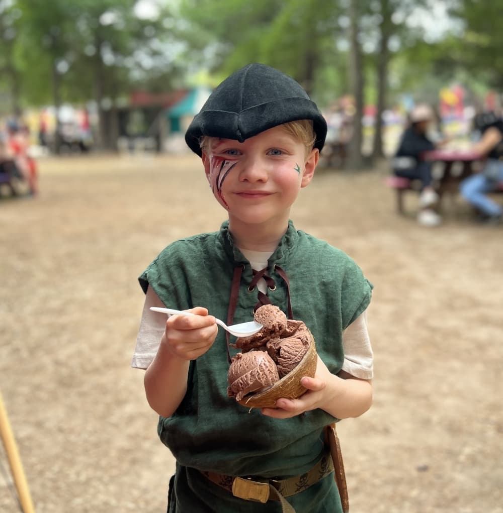 A small child dressed in medieval costume eats ice cream out of a coconut shell.