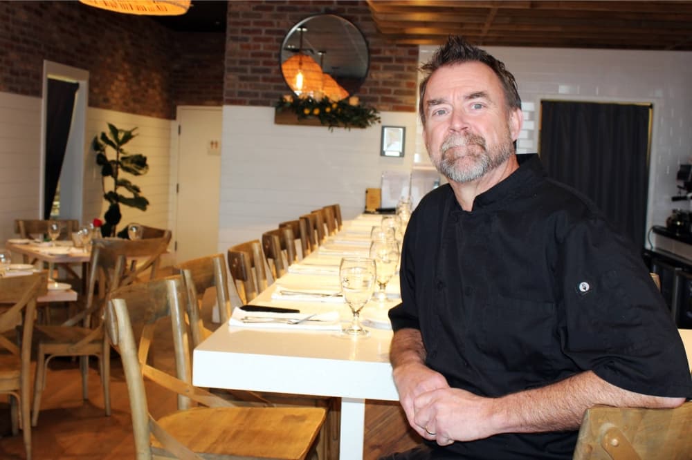 Male wearing a black chef jacket sitting at bar.