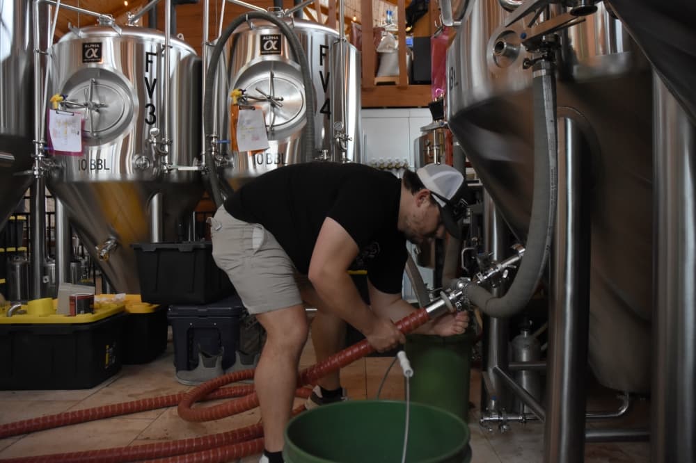A man bends over to attach a hose to a metal tank in a brewery. 