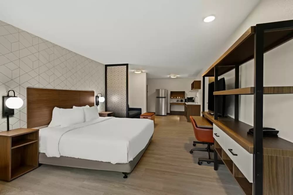 Inside a room is shown, with a bed and entertainment center in the foreground, and a kitchenette in the background.