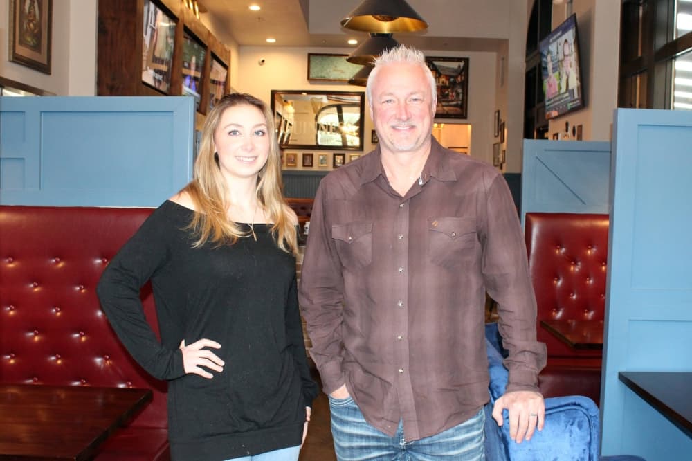 Female and male standing side-by-side inside a restaurant with steel blue wooden booths padded in red tufted leather. 