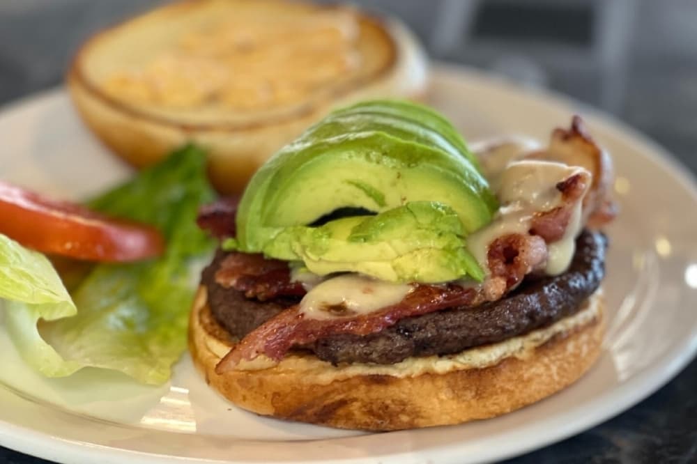 Burger topped with avocado and cheese with a side of lettuce and tomato. 