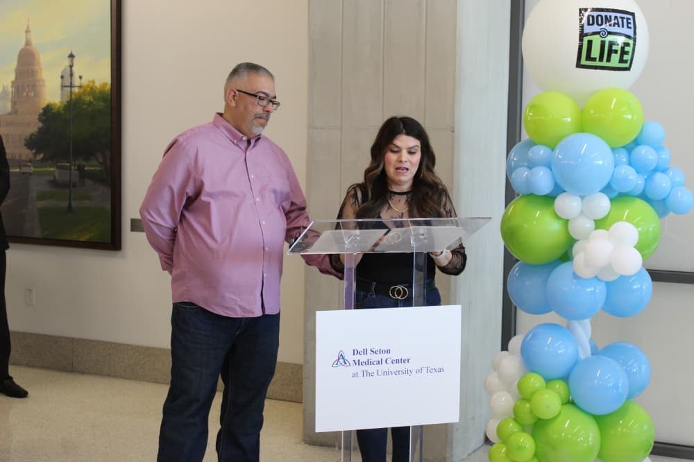 A man stands next to his wife who speaks at a podium