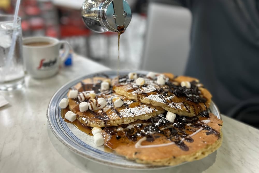 Person pouring syrum on a stack of pancakes topped with chocolate sauce. 