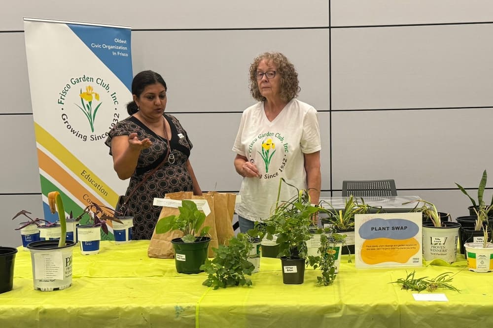 Two women standing behind a table with various plants sitting on it. 