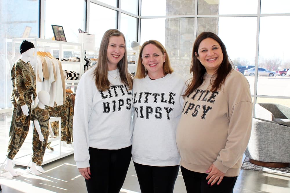 Three women wearing Little Bipsy branded sweatshirts. 