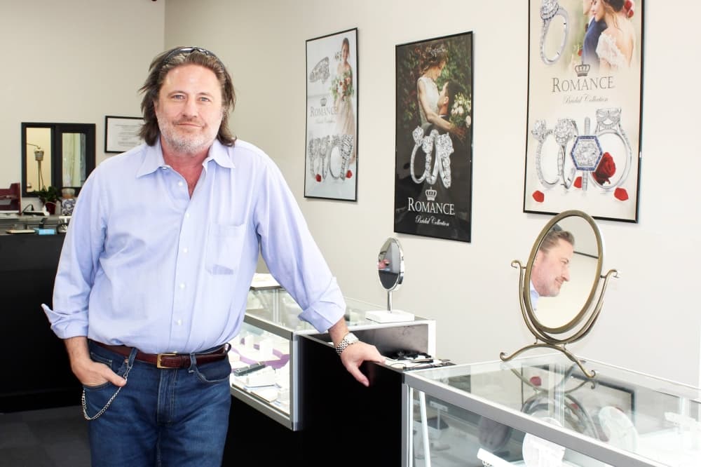 Male standing near a jewelry store case with one hand on the case, the other hand in his pocket. 