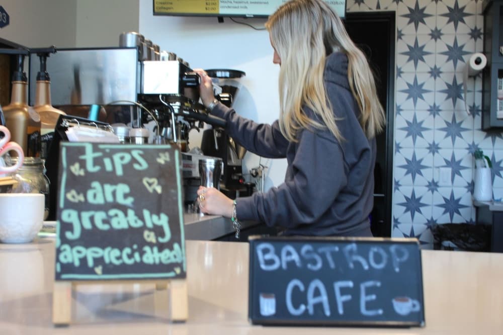 An employee makes coffee in the background which is in focus, with Bastrop Cafe signs on the counter that are blurred in the foreground.