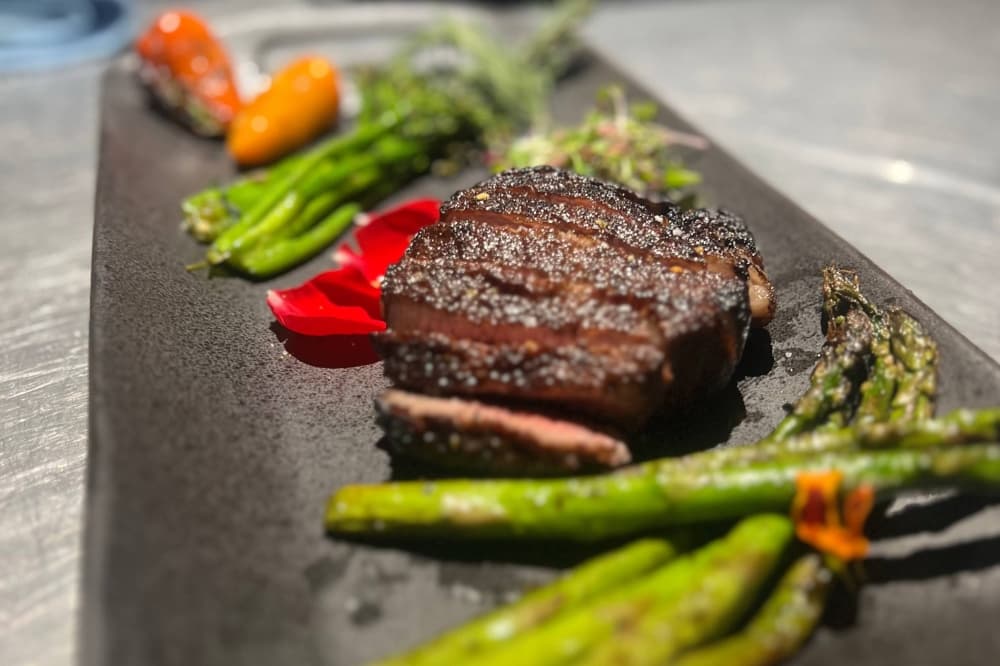 Filet mignon on a dark rectangular plate flanked with asparagus and other vegetables. 
