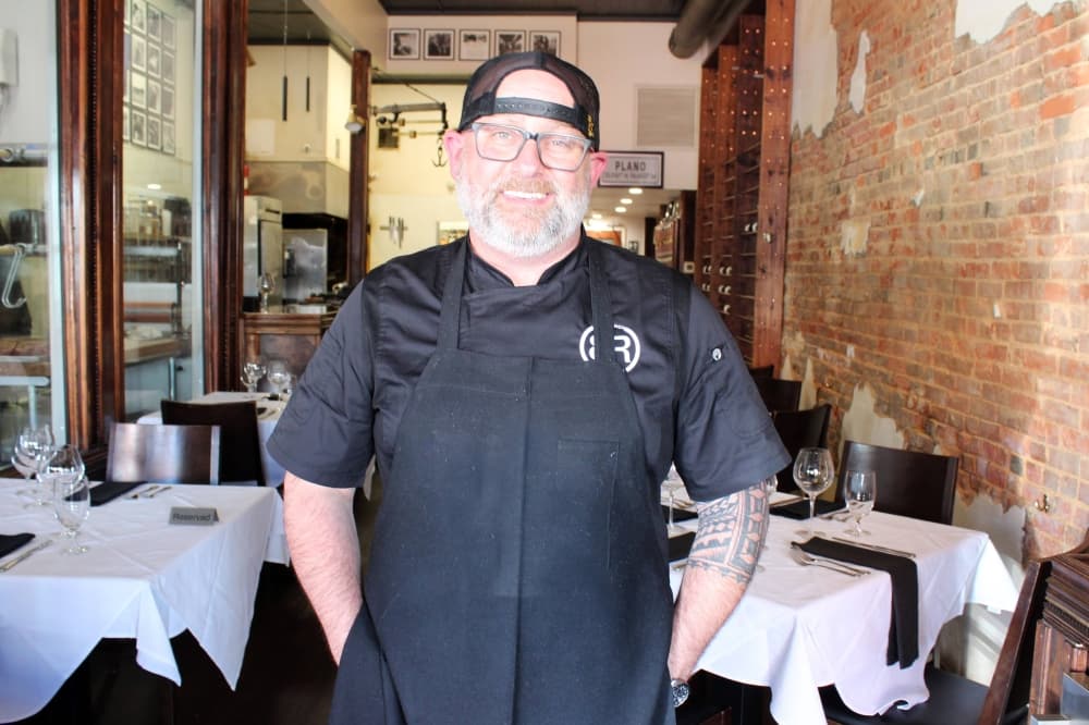 Male wearing black apron, shirt and ballcap standing in a restaurant with tables topped with white cloth tablecloths. 
