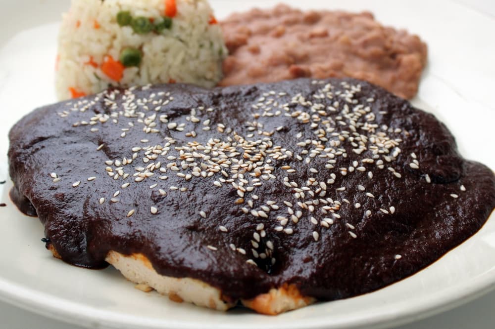 Chicken on a plate topped with mole sauce with sides of rice and beans. 