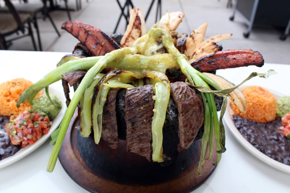 Large lava bowl filled with meat and vegetables with sides of rice and beans. 