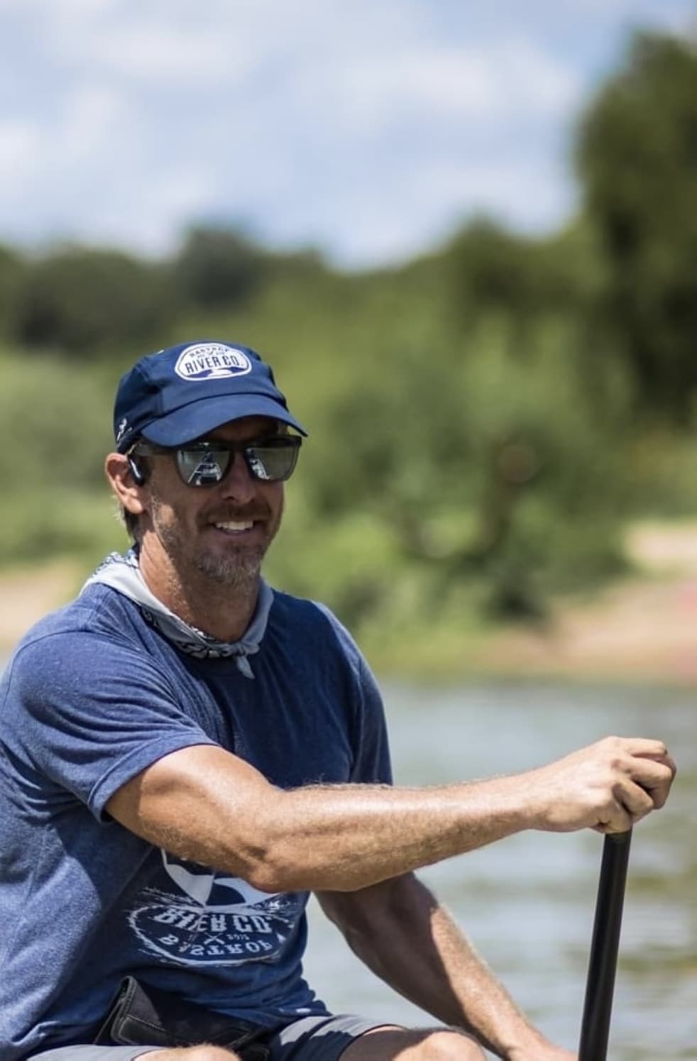 A tight shot of the owner paddling down a river and smiling while wearing a cap and sunglasses.
