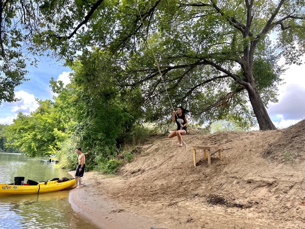 Teenagers are shown along the bank near a kayak, one is swinging from a rope swing tied to a large green tree.