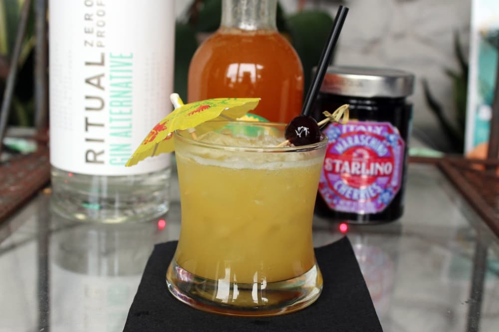 Yellow drink garnished with paper umbrella sitting in front of dry bar ingredients. 