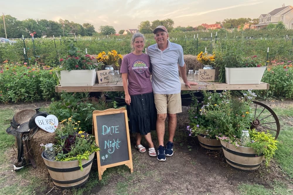 Female and male standing next to a cart in front of a crop of blackberry plants. 