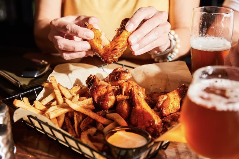 a woman holds a chicken wing over a basket of fries