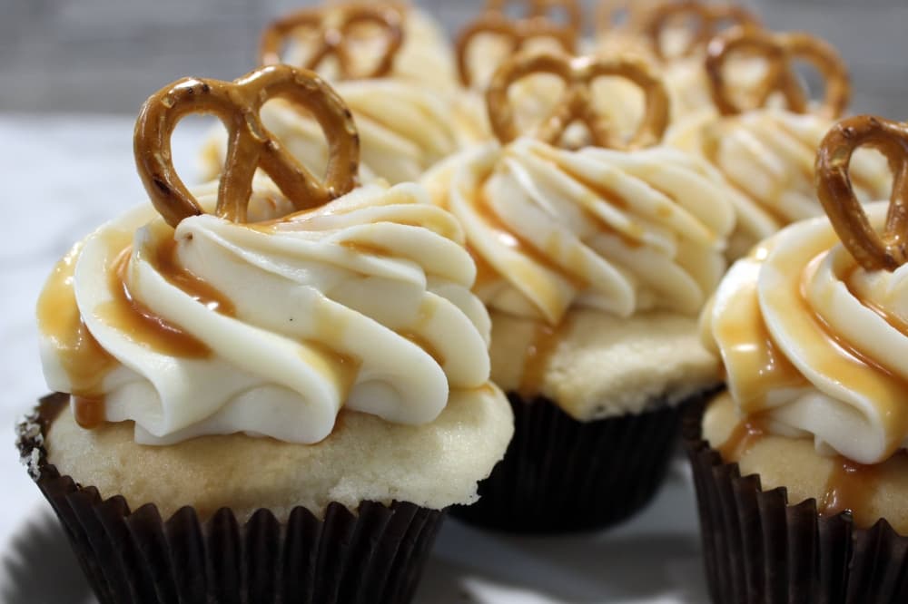 Cupcakes topped with white icing drizzled with caramel sauce and topped with a pretzel. 