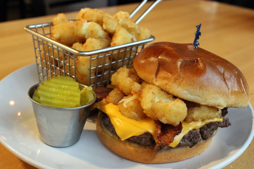 Cheeseburger topped with fried cheese curds with a side of cheese curds and pickles. 