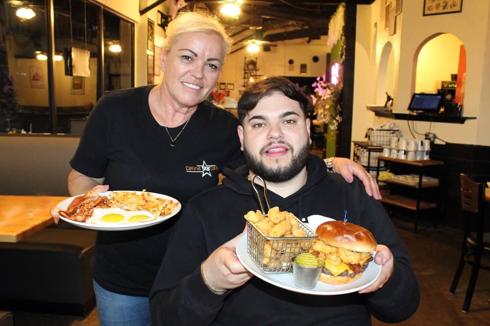 Female standing next to a seated male in a restaurant. Both people are holding plates of food. 