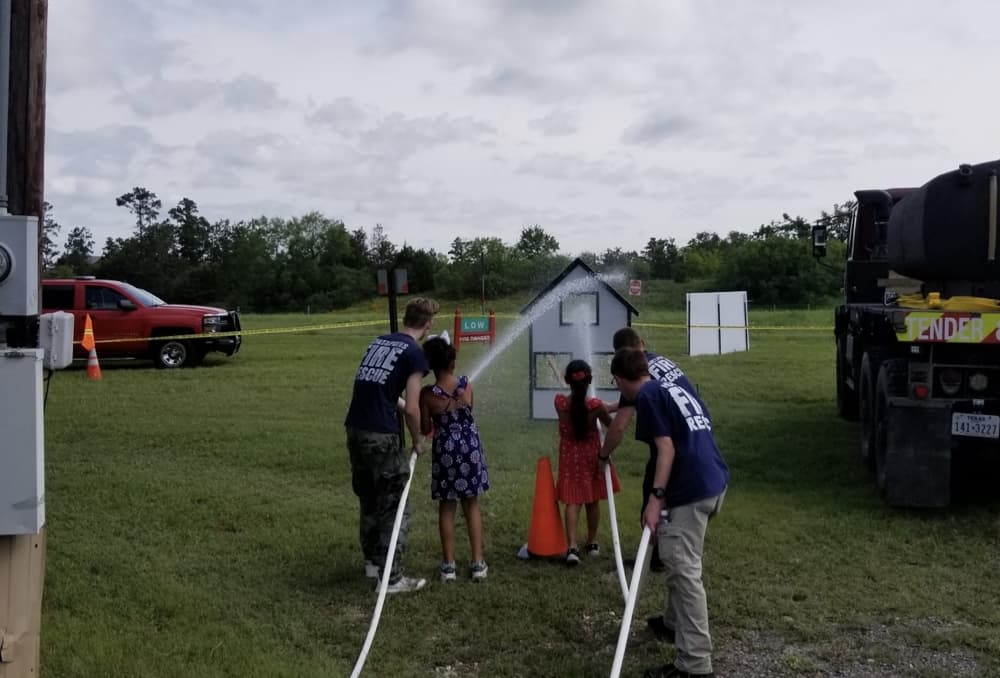 An image of two firefighters helping children spray a firehose. 