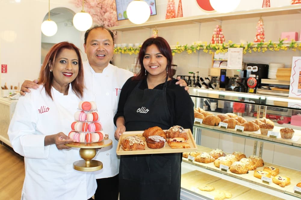 Two females and a male standing together in a bake shop in front of a full bakery case. 