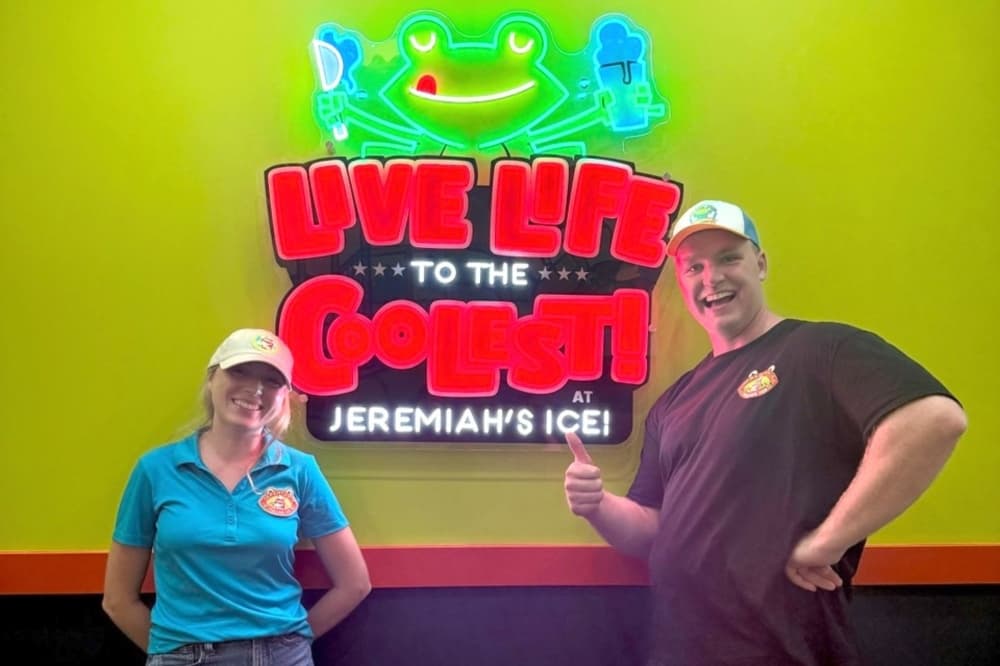 The couple wearing official shirts and hats stand in front of the neon sign inside the Bastrop location for the company. Both smiling, Jack also gives a thumbs-up.