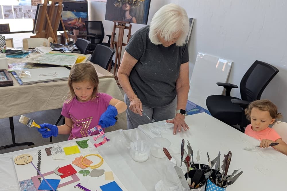 Two children seated, and one adult standing, at tables working on art projects.