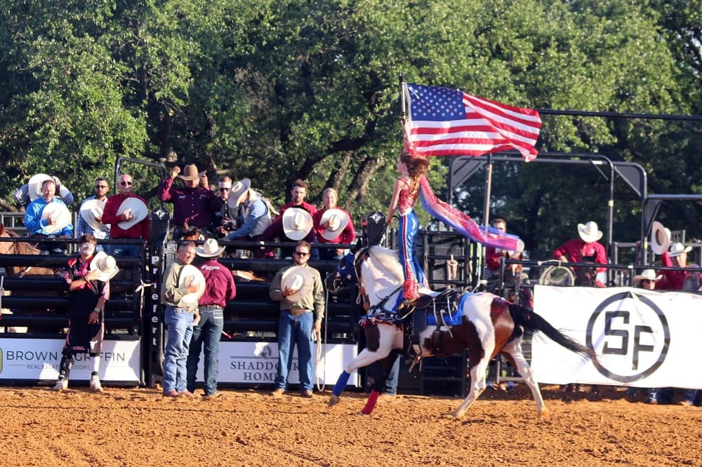Female standing on a horse while holding an American flag and going around a rodeo arena. 