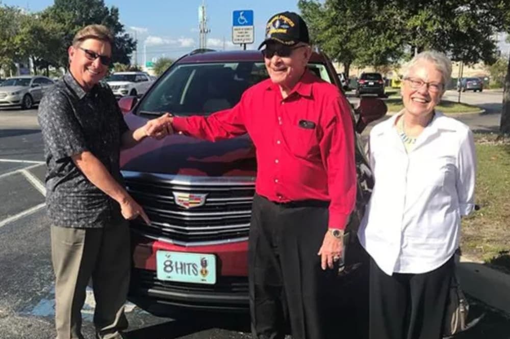 JohnnySanders, Billy Waugh and his wife Lynn Waugh laugh in front of Waugh's vehicle, which has a license plate that reads "8 hits" to symbolize his 8 purple hearts.