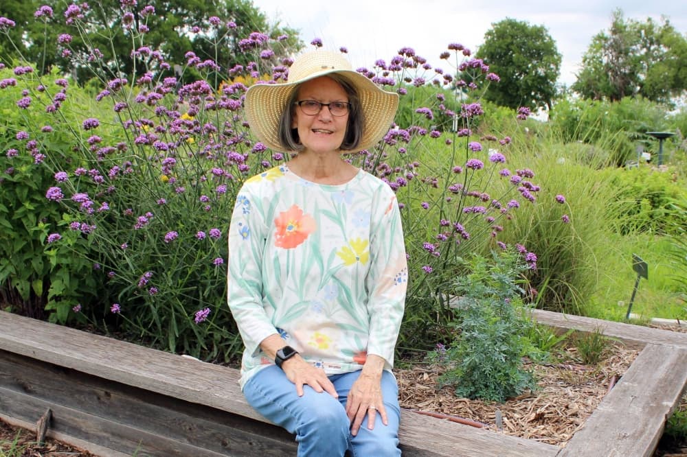 Female sitting on a raised flower bed with a purple-bloom plant behind her. 