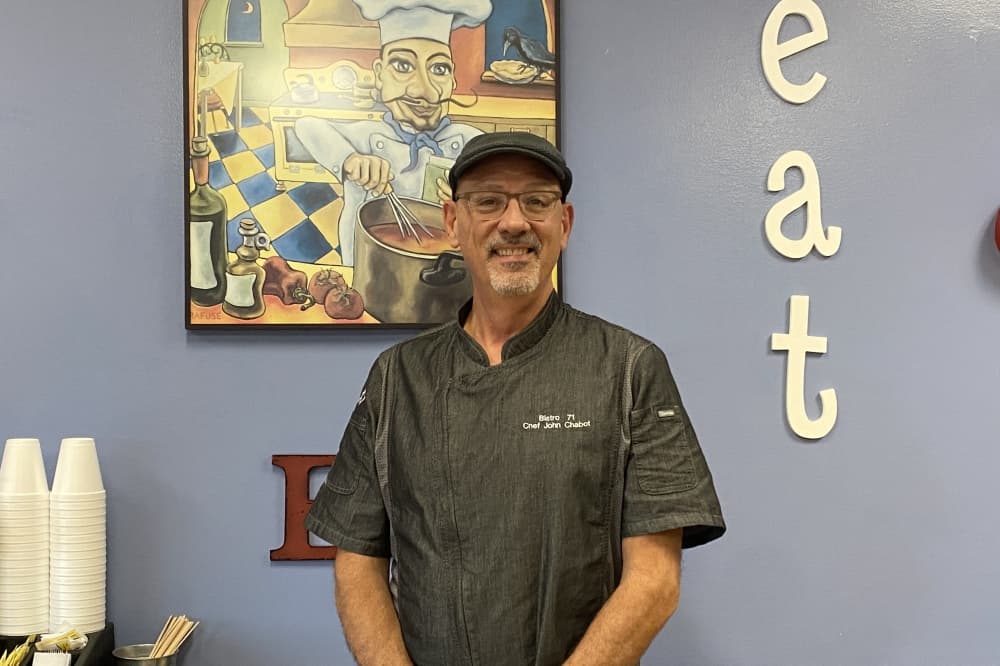 Chef stands in front of a blue restaurant wall. He wears a monogrammed chefs coat, a hat and glasses. To his right, there are wall letters that say eat and behind him is a big picture of a cartoon chef. 