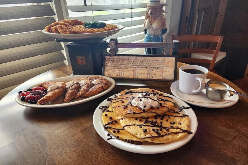 A plate of pancakes topped with chocolate chips, a fresh cup of coffee, a plate of fruit-covered croissants, and a dish of chicken & waffles.