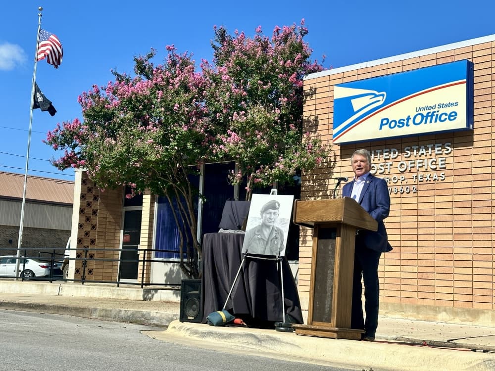 Wearing a suite, McCaul speaks in front of the post office adjacent to a large photo of Billy Waugh with the US and POW flags in the background.