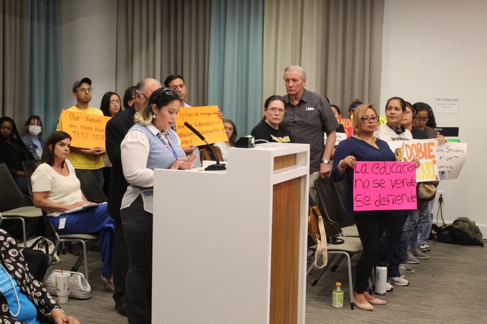 A woman speaks at the podium at a school board meeting next to people holding signs