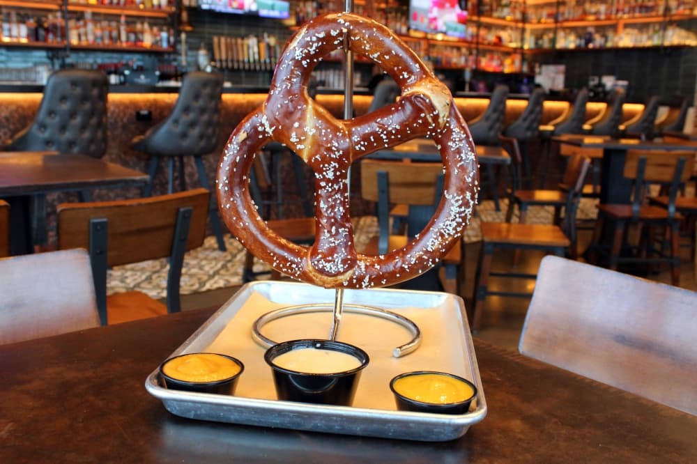 Large pretzel on a metal pole mounted on a silver tray with small bowls of dipping sauce. 