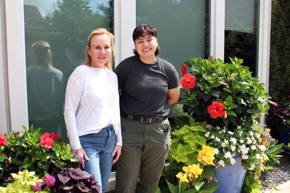 Two females standing next to each other between urns filled with flowers. 