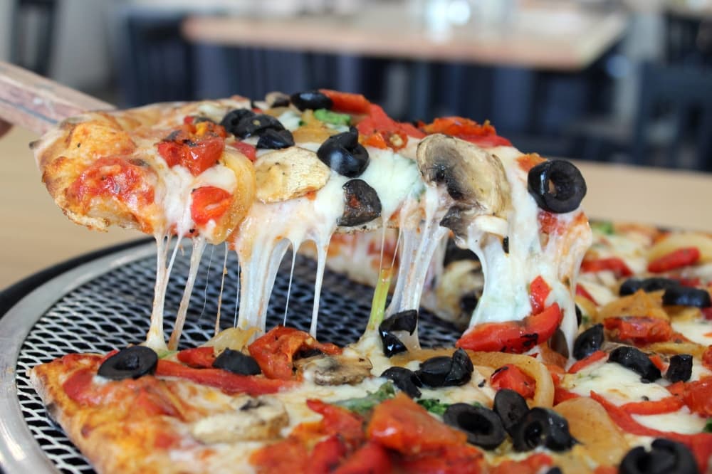 One slice of veggie pizza being pulled up from a pizza pie while strands of melted cheese connect the items. 