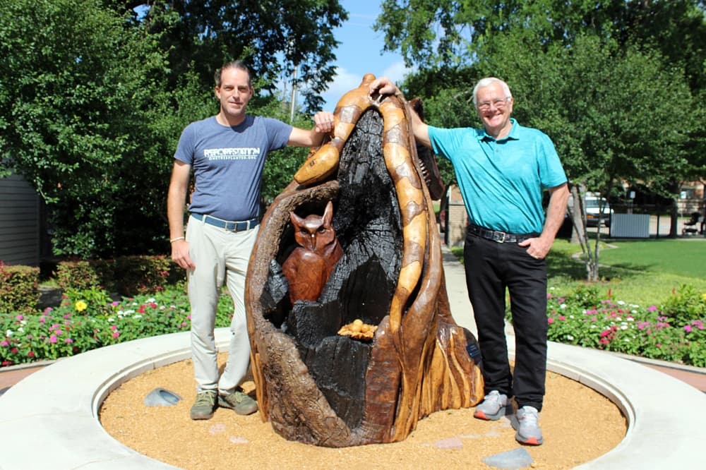 Two adult males standing on either side of a tall wooden sculpture of animals displayed outside. 