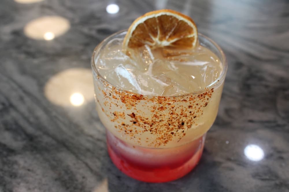 Cocktail glass filled with light-colored drink with a rim of red-toned salt. 