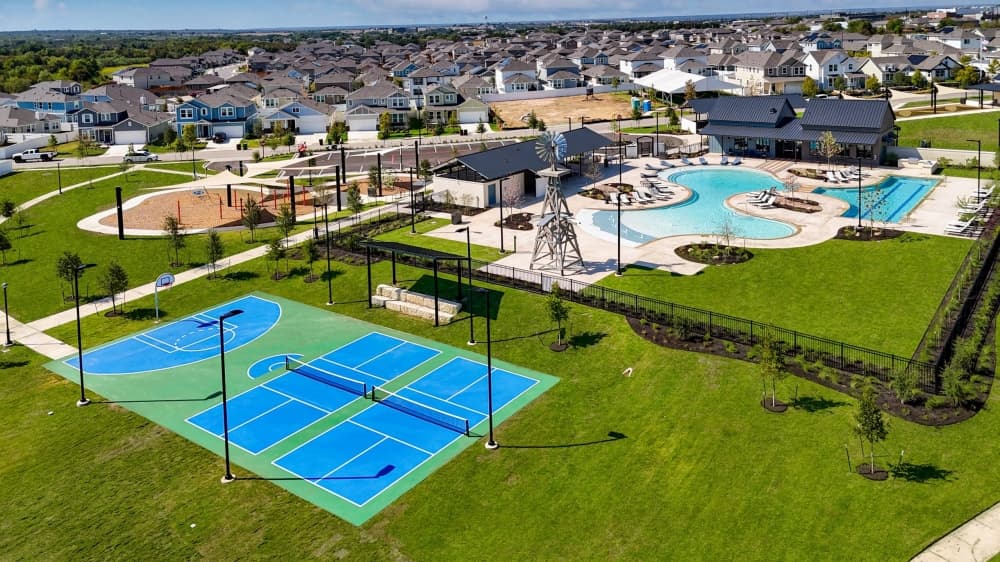 Aerial view of a Brohn Homes master-planned community featuring a resort-style pool, modern clubhouse, basketball court, pickleball courts, playground, and landscaped green spaces surrounded by newly built homes