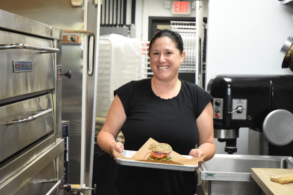 A woman stands in a commercial kitchen holding a bagel sandwich. 
