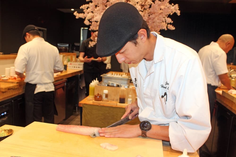 Sushi chef wearing white chef jacket and black cap while slicing fish at a sushi bar station. 