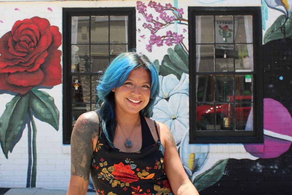 Female with blue and black hair sitting in front of the exterior of a business featuring a flower mural. 