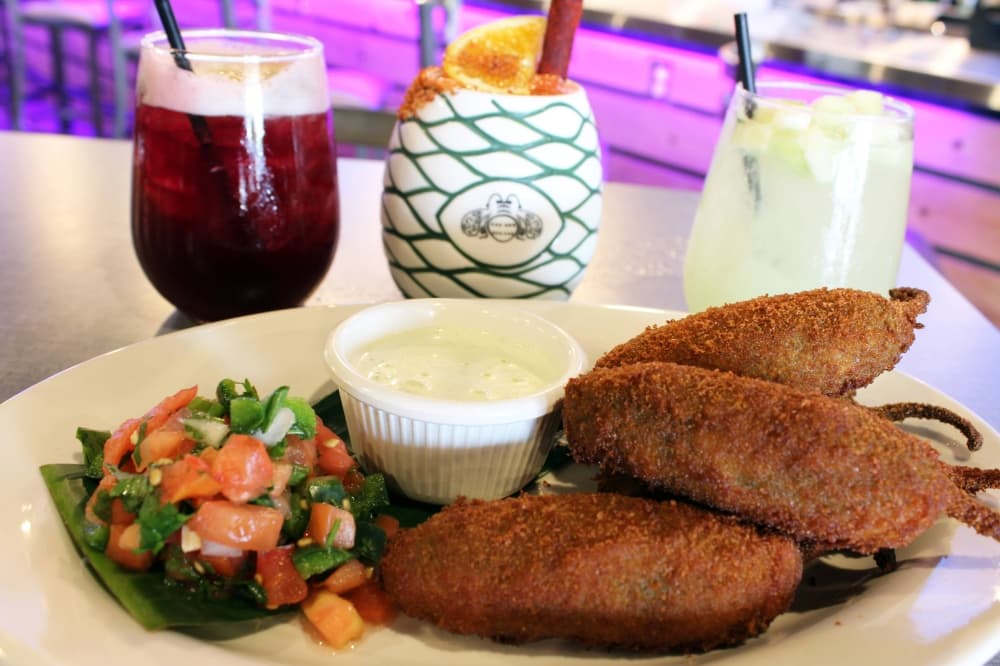 Five jalapeno relleno on a plate with three drinks in the background. 