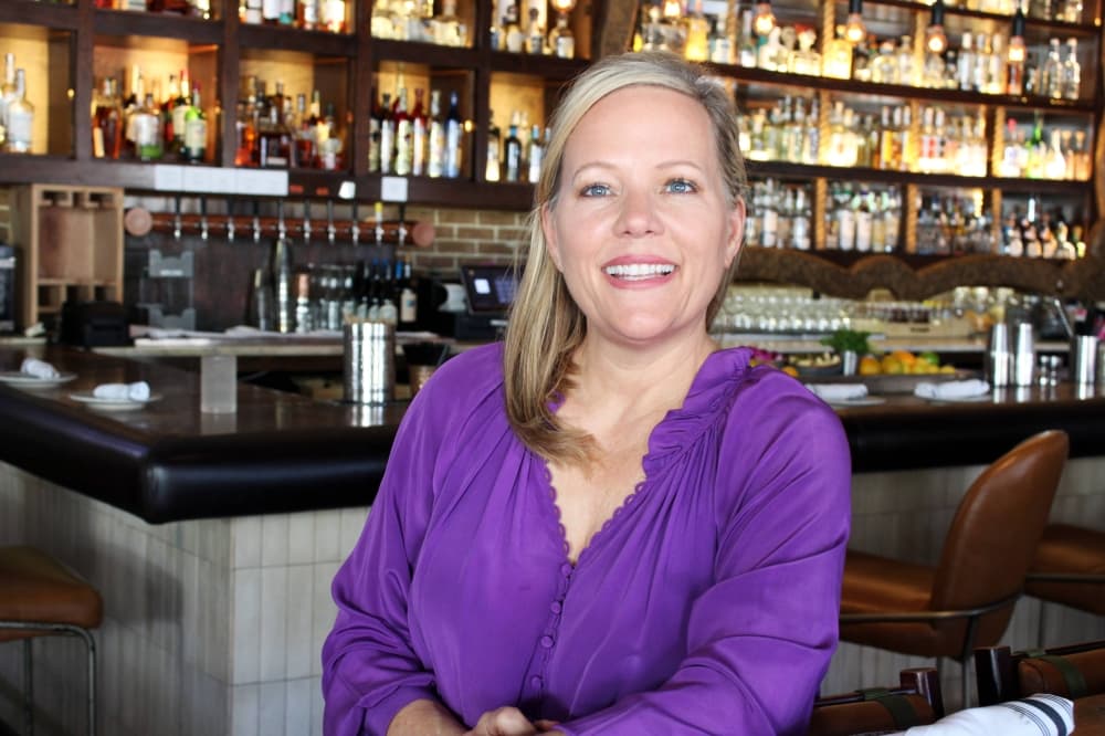 Female leaning against a counter in a restaurant with a bar behind her. 