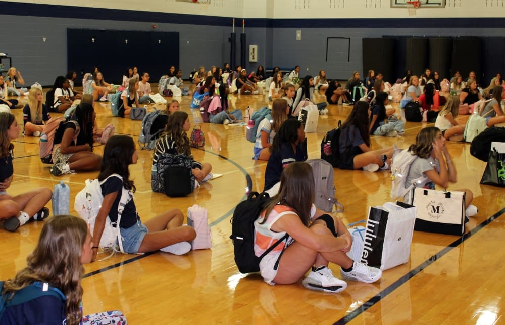 Female teens sitting on an indoor basketball court. 